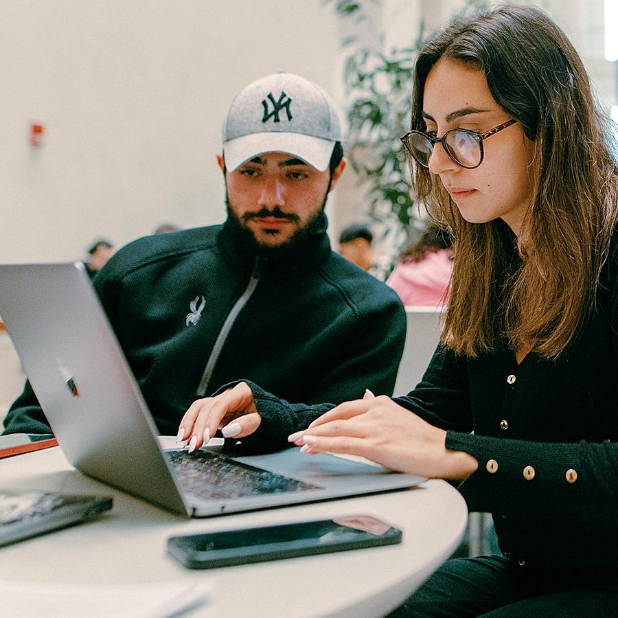 Two students one in baseball hat look over laptop in campus center.